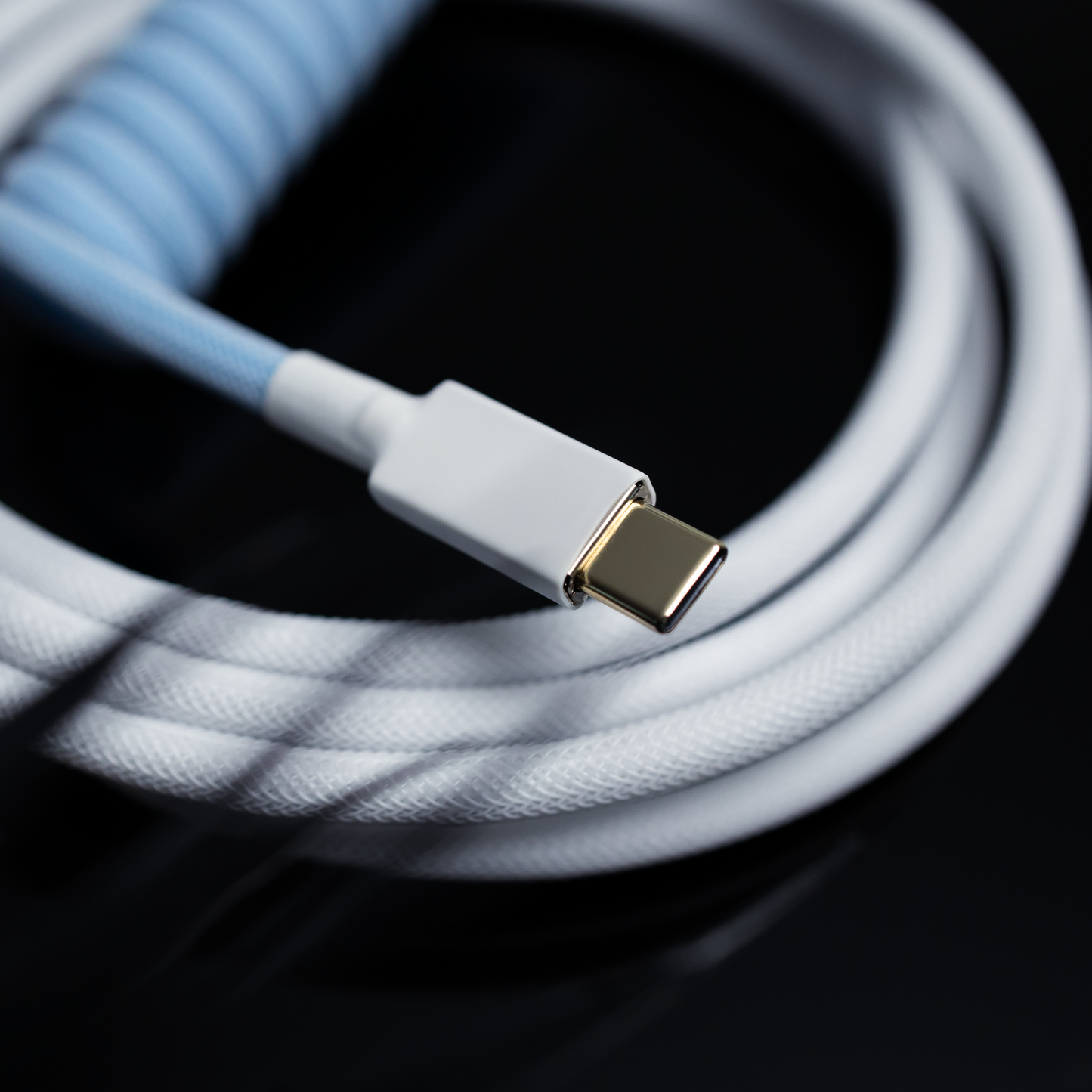 Closeup of gold USB C with white heat-shrink. In the background, a coiled cable with Aqua Blue Teleios, a straight cable with Polar White Teleios, and white TechFlex.
