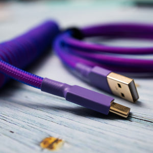 Closeup of USB C connector with purple heat-shrink tubing. In the background, a gold USB A connector with purple heat-shrink tubing and the rest of the mechanical keyboard cable exhibiting pink Teleios sleeving and blue TechFlex  
