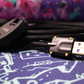 Closeup of CNC USB A connector with purple PCB and wrapped black on black sleeved Teleios mechanical keyboard cable with a Canon camera lens out of focus on a multi colored desk mat. 