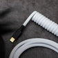 Closeup of white and black coiled mechanical keyboard cable. Teleios Polar White sleeving, white TechFlex, black heat-shrink tubing, gold USB A and USB C connectors, black sandblasted FEMO 1B push/pull connector.