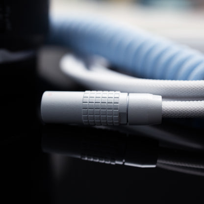 Closeup of white sandblasted FEMO 1B (female end). In the background, the coiled end of the cable in Teleios Aqua Blue, the straight end in Teleios Polar White, and white TechFlex.