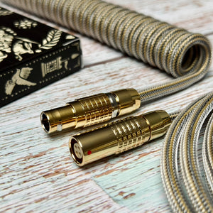 Closeup of gold FEMO 1B connectors and coiled mechanical keyboard cable with Teleios Fusion Black and Gold sleeving, clear TechFlex second later, and a deck of black and gold playing cards just out of frame. 