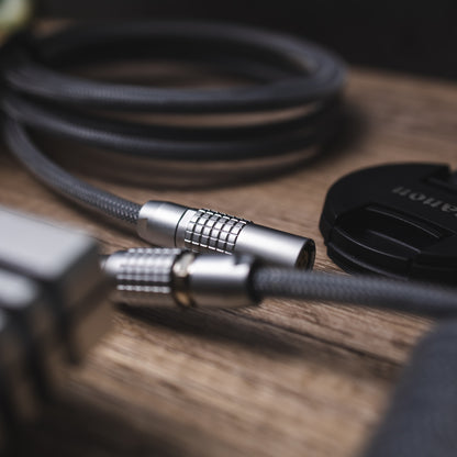 Closeup of FEMO female connector. A canon lens cap half out of frame. Teleios carbon grey/gray, TechFlex Clear, FEMO 1B push/pull connector, and bundled cable of the same type. 